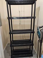 Black hard plastic shelving unit with five slatted shelves and solid frame standing empty against a wall