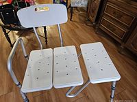 Photo shows a white plastic three-section shower seat with a metal frame. Two legs extend inside the bath area, and two legs extend outside the tub. The seat has ventilation holes and an attached backrest with a handle opening.