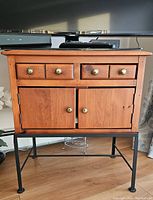 Front view of wooden table showing four drawers and two cabinet doors with gold-colored knobs, black metal legs on wooden floor.