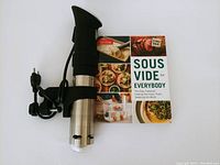 Anova sous vide precision cooker with power cord and accompanying cookbook 'Sous Vide for Everybody'. The cooker is stainless steel and black plastic with clamp attachment visible.