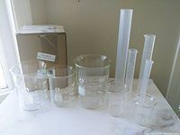 Set of nine graduated cylinders including five glass cylinders with white measurement markings and four translucent plastic cylinders. Items arranged on a flat surface near a window.