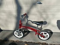 Side view of red Radio Flyer push bike with white plastic tires and black seat, shown outdoors in daylight.