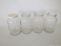 Front view of four clear glass jars in a row; one jar has a white lid, three jars have no lids.