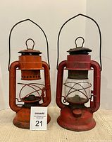 Pair of antique kerosene lanterns showing front sides with handles and ventilation top, metal chips and rust visible on older red paint.
