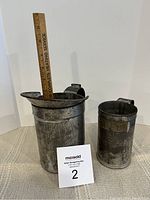 Two antique tin measuring cups side by side showing overall size and condition. Larger cup has metal handle and spout, smaller cup is marked 'PINT'. A wooden ruler indicates the larger cup height.