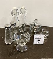Top-down angled photo showing all glassware items including bottles, shakers, bowls, creamer, small pitcher and small cup with lot number card