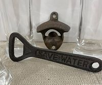 Close-up image showing hand-held cast metal bottle opener with embossed text "SAVE WATER" in front of wall-mounted cast metal O'Keefe bottle opener and two clear beer glasses.