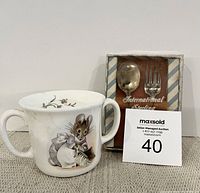 Bone china baby cup showing Beatrix Potter mouse character on side with two handles and floral decoration inside rim. Also shows boxed International Sterling baby spoon and fork set behind cup.