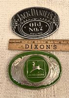 Two belt buckles positioned side by side with a ruler for scale. One buckle features Jack Daniel's logo in black and silver, the other features John Deere logo in green and silver.