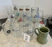 Photo showing multiple branded beer glasses and mugs arranged on a textile surface, including variety of tall and short glasses with logos such as Kilkenny, Stella Artois, Hoegaarden, Smithwick's, Rickard's, and a green Alexander Keith's ceramic mug.