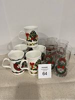 Overview of six white ceramic Christmas-themed mugs alongside ten clear glasses decorated with wreath and red bow motifs