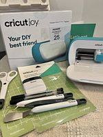 Photo of Cricut Joy machine, box, and assorted accessories including scissors, pens, scraper, and mats spread out