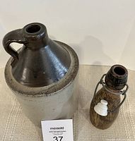 Two crockery jugs, large one with beige lower portion and dark brown top including handle and spout, smaller jug mottled brown with white ceramic stopper and wire clasp.