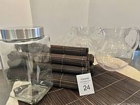 Photo showing glass lidded jar with metal lid, clear glass pitchers, and stacked dark brown bamboo placemats and napkin holders.