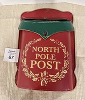 Front view of mailbox showing red body with gold 'NORTH POLE POST' text surrounded by a holly wreath, and green flap at top.