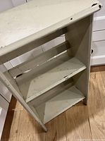 Front view of white painted wooden side table/book rack showing two shelves and top surface, with visible paint wear and small chips.