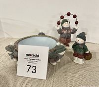 Photo of the three snowmen figures on beige surface with MaxSold auction card.