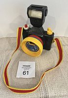 Front view of vintage Fisher Price Crazy Camera toy with colored lens ring, flash piece on top, and red-yellow strap.