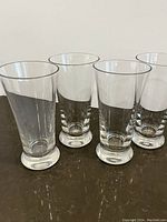 Four clear vintage drinking glasses arranged on a dark surface, showing tapered cylindrical shape and thick bases.