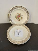 Two vintage luncheon plates shown, one with a basket of colorful flowers in the center, the other with an intricate Century gold pattern around the rim. Both have 23k gold trim.