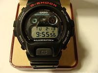 Front view of the Casio G-Shock DW-6900 digital watch showing the time, day, and date on the negatively displayed screen with the G-Shock branding in red above and button labels on bezel.