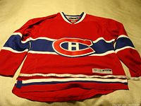 Full front view of red Montreal Canadiens hockey jersey with measurement tape showing size, displayed flat on beige textured surface