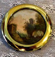 Top view showing the detailed watercolor landscape painting inside shiny brass circular frame on lace tablecloth