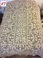 Full view of cream-colored handmade lace tablecloth laid over a bed, showing intricate floral and leaf lace patterns and scalloped edges.