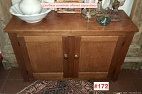 Front view of the antique pine cupboard showing two paneled doors and wooden knobs, resting on a decorative rug with additional items on top.