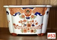 Front view of the rectangular white pottery plant pot showing detailed floral and geometric painted designs in orange, blue, green, and beige. The pot has small feet and a flared rim.