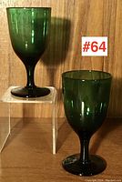 Pair of green Victorian hand blown wine glasses displayed against a wood background, with one elevated on a clear stand.