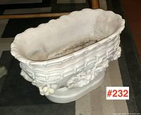Front angle of the vintage white ceramic planter on a carpeted floor with visible fruit and leaf relief pattern around the sides and scalloped top edge.