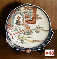Front view of fan-shaped vintage Asian pottery serving dish showing detailed painted scene with table, mythical creatures, flowers, and teapot.