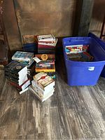 Photo of stacked adult fiction books including thick paperbacks and hardcovers next to a blue plastic tote containing children's books