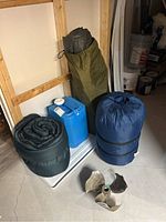 Full lot showing all items including two sleeping bags, tent bag, water jug, and boat propeller on basement floor.