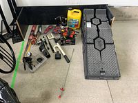Overview photo showing Rhino ramps, battery charger, battery cables, wiper blade, trailer hitch, paint sprayer, and yellow automotive fluid container on garage floor.