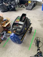 View of the full air compressor unit showing the blue tank, black casing, wheels, and surrounding garage environment.