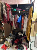 Wide view of an open wardrobe filled with various costumes including shirts, jackets, a gold dress hanging on the door, bins and piles of assorted fabrics and costume items on the floor beneath.