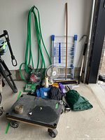 Photo showing entire lot including the four wheeled movers dolly, green garden hose, bike lock, collapsible clothes dryer, Comfort Zone fan, blue tarp, green rain poncho, push broom, and other items arranged on garage floor and wall.