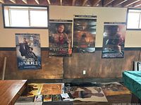 Four large movie posters mounted on the wall: Tomb Raider, Titanic, Godzilla, and The Last of the Mohicans. Several smaller posters or playbills are laid flat on the table beneath.