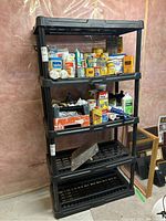 Front view of the black resin shelving unit containing various household items like paint cans and bottles, showing four shelves with slats and sturdy plastic construction.