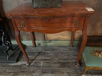 Front view of a wooden table with single drawer and metal pulls on curved legs.