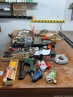 Wide shot of lot showing wooden table with assorted tools and accessories spread out, including pneumatic brad nailer, tape measures, drill bits, screwdrivers, and other small hand tools.