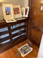 Corner display of multiple matted and loose unframed artworks along a wooden shelf, plus prints on the floor.