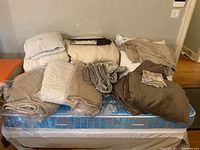Full collection of bedding items arranged on mattress, including duvets, blankets, pillows, and fabric pieces in neutral tones.