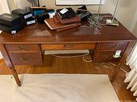 Wood desk with five drawers under a protective clear mat, medium brown finish, located indoors on a light carpet, with various items on top that are not included.