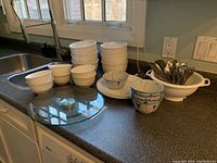 View of six large white bowls stacked, multiple small textured white bowls, glass lazy Susan, Dansk Odin cutlery in white strainer on kitchen counter.