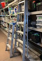 Two metal step ladders side by side in a storage area with shelves holding various items.