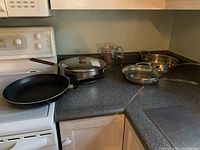 Wide view of the kitchen countertop showing two frying pans (one with lid), two Pyrex measuring cups, and another frying pan with lid and handle.