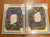 Two framed geometric prints side by side on wood floor with protective corner tape and auction tag.